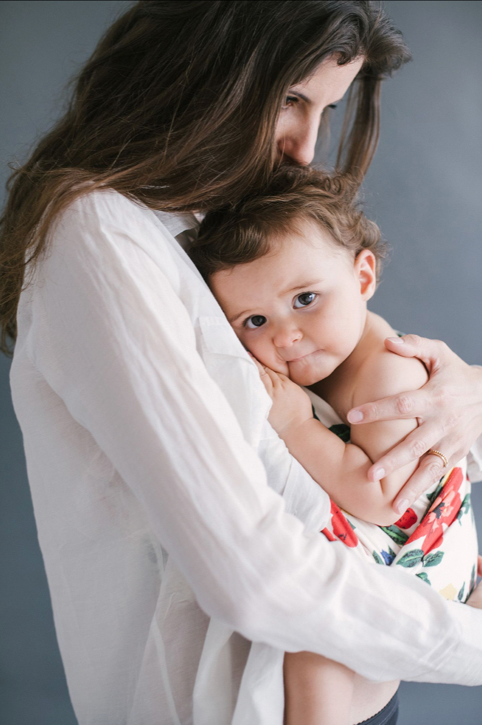 A long-haired mother tenderly embraces her baby in a floral outfit against a gray background.