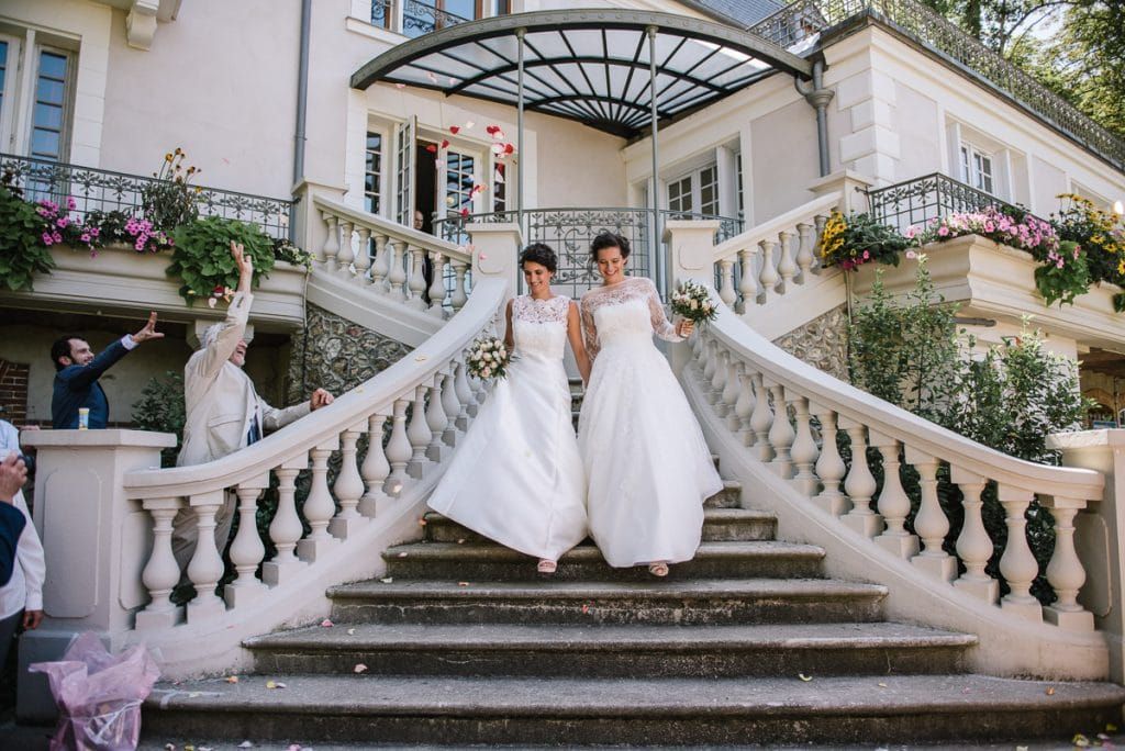 Two brides in white gowns descend grand stone staircase of elegant French chateau on their wedding day.