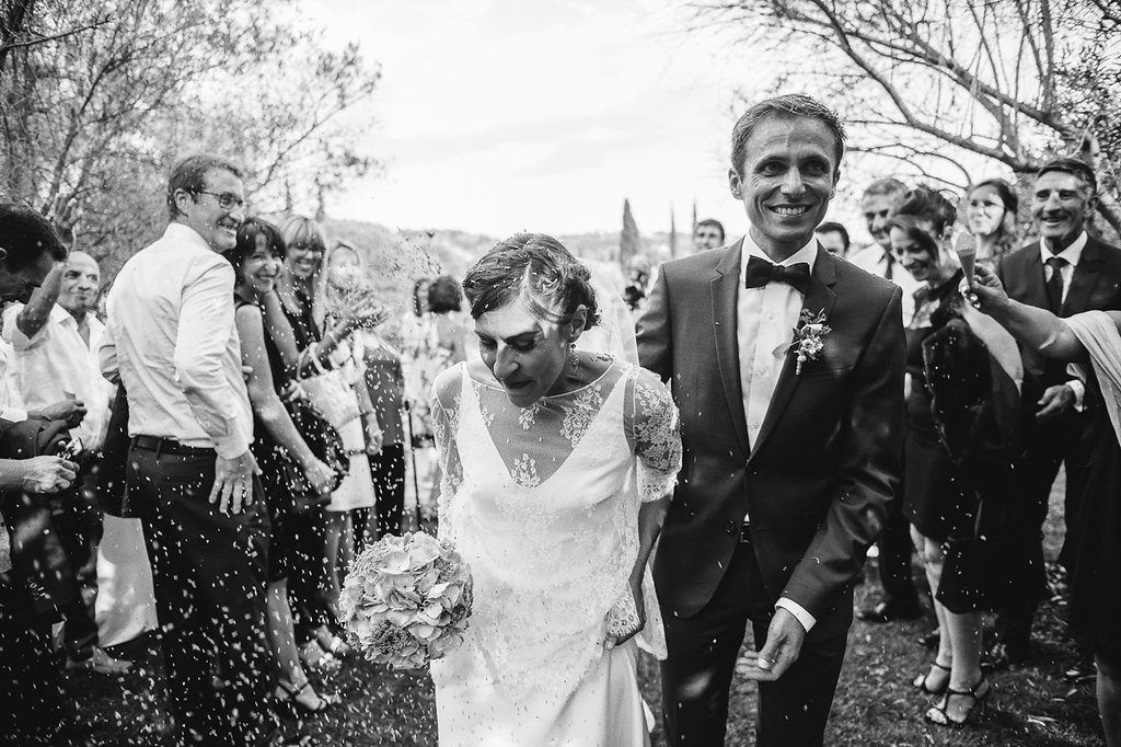 Black and white photo of smiling groom leading bride through confetti shower among celebrating wedding guests.