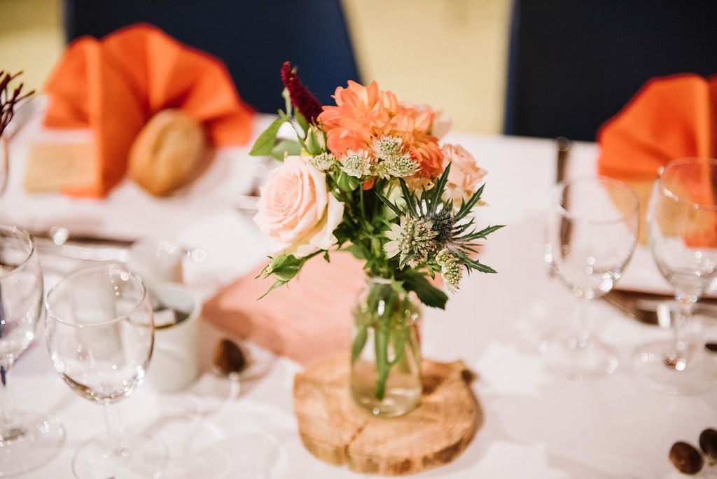 Elegant wedding table centerpiece with peach and pink flowers in a glass jar on a rustic wood slice.