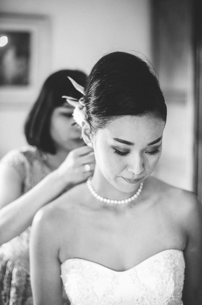 Assistant placing earring on bride wearing pearl necklace and strapless lace gown.