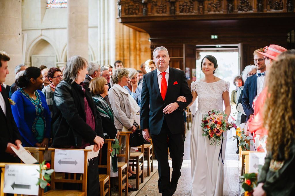 Bride walking down church aisle with father in red tie holding colorful bouquet.