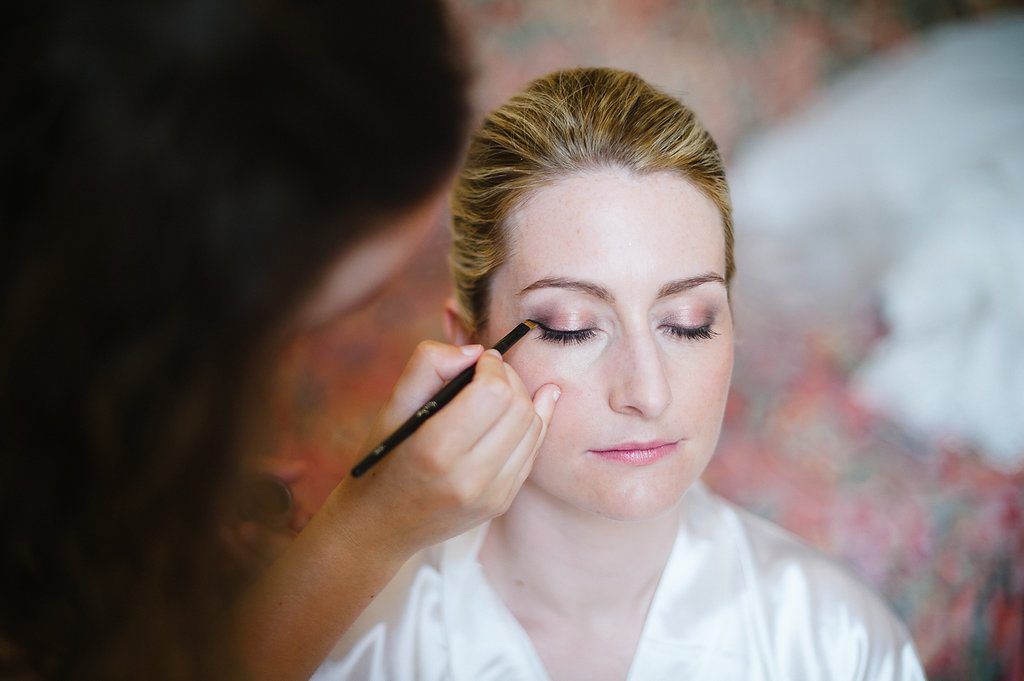 Makeup artist applying eye makeup to a blonde bride in a white satin robe.