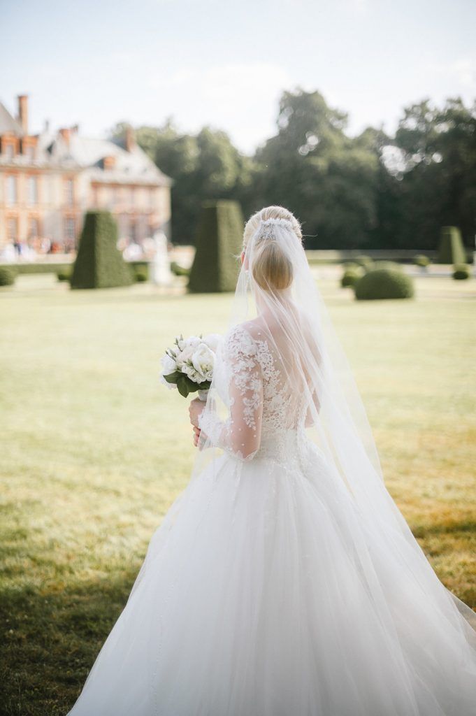 Bride in lace wedding dress with veil holding bouquet before French chateau garden.