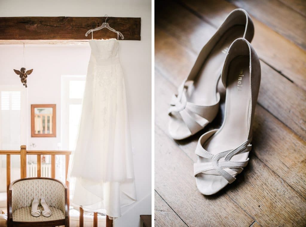 White lace wedding gown hanging near window beside elegant white open-toe bridal heels on wood floor.