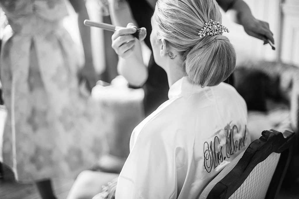 Black and white photo of bride in monogrammed robe getting makeup applied before wedding.