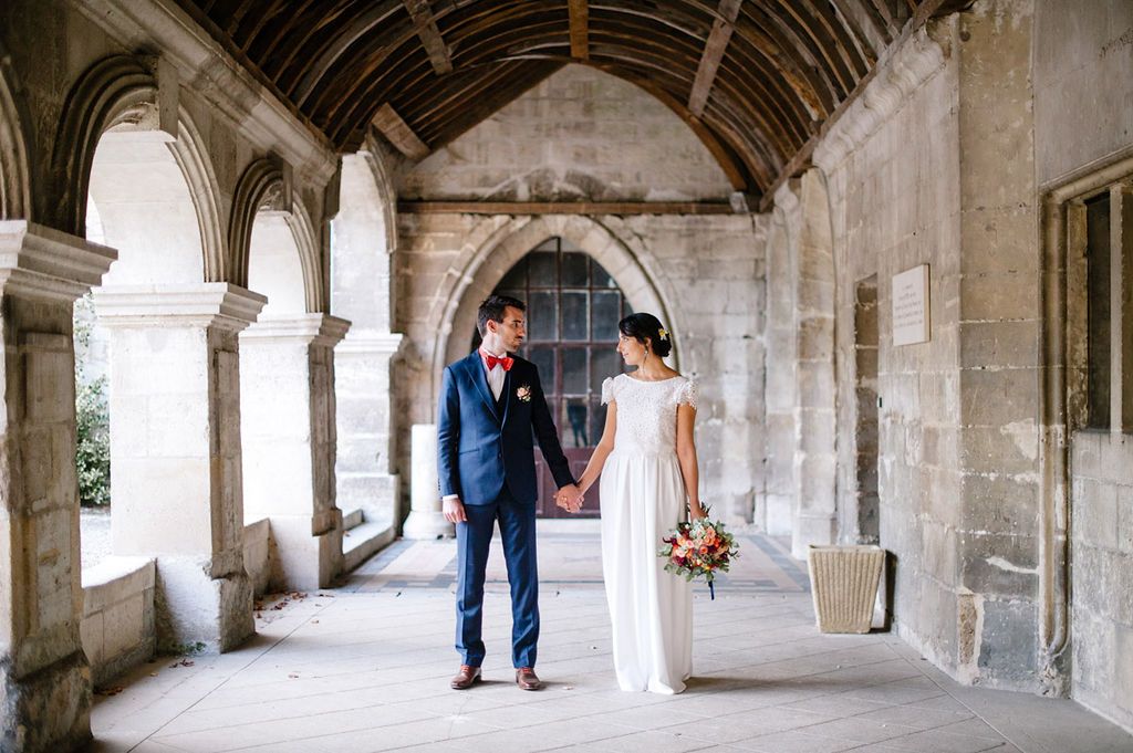 Bride and groom hold hands in elegant stone archway corridor of historic wedding venue.