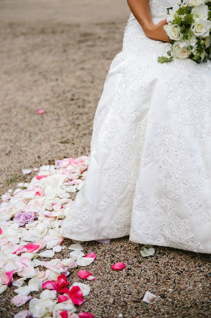 Bride in embroidered white lace gown holds bouquet standing over scattered pink and white rose petals.