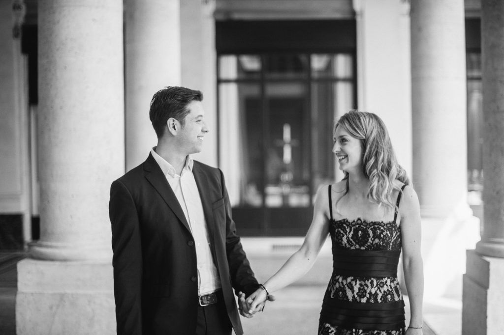 Black and white photo of a couple holding hands and smiling at each other between elegant white columns.