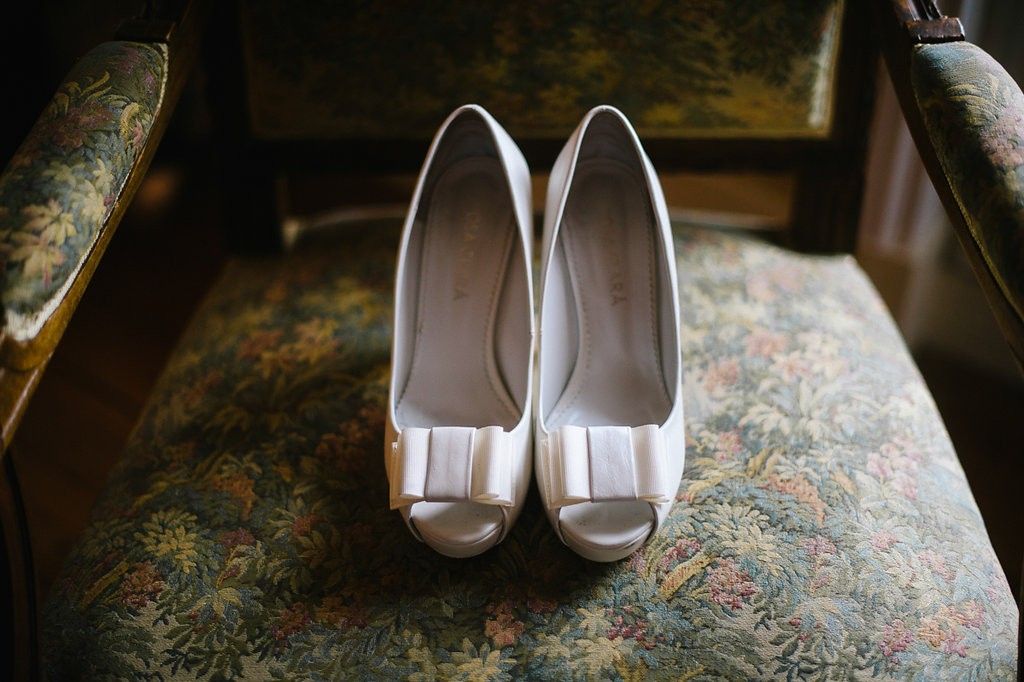 White peep-toe bridal heels with bow detail resting on a vintage floral upholstered chair.