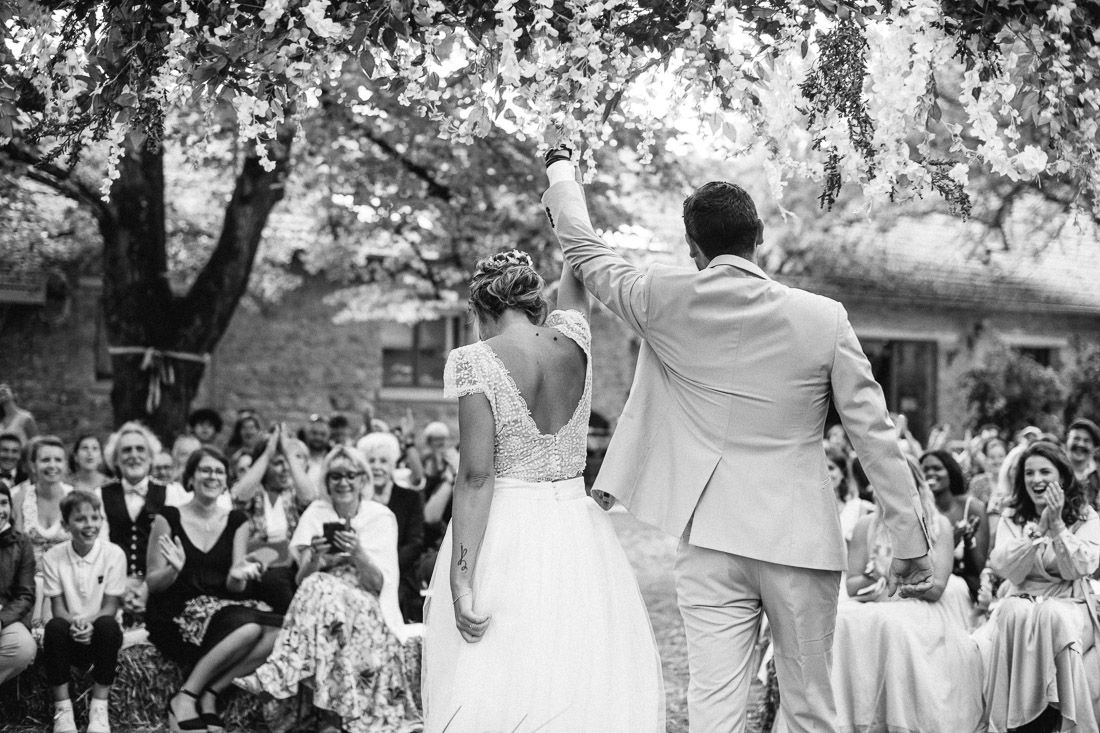 Newlyweds celebrate joyfully under flowering tree as wedding guests cheer in black and white photo.