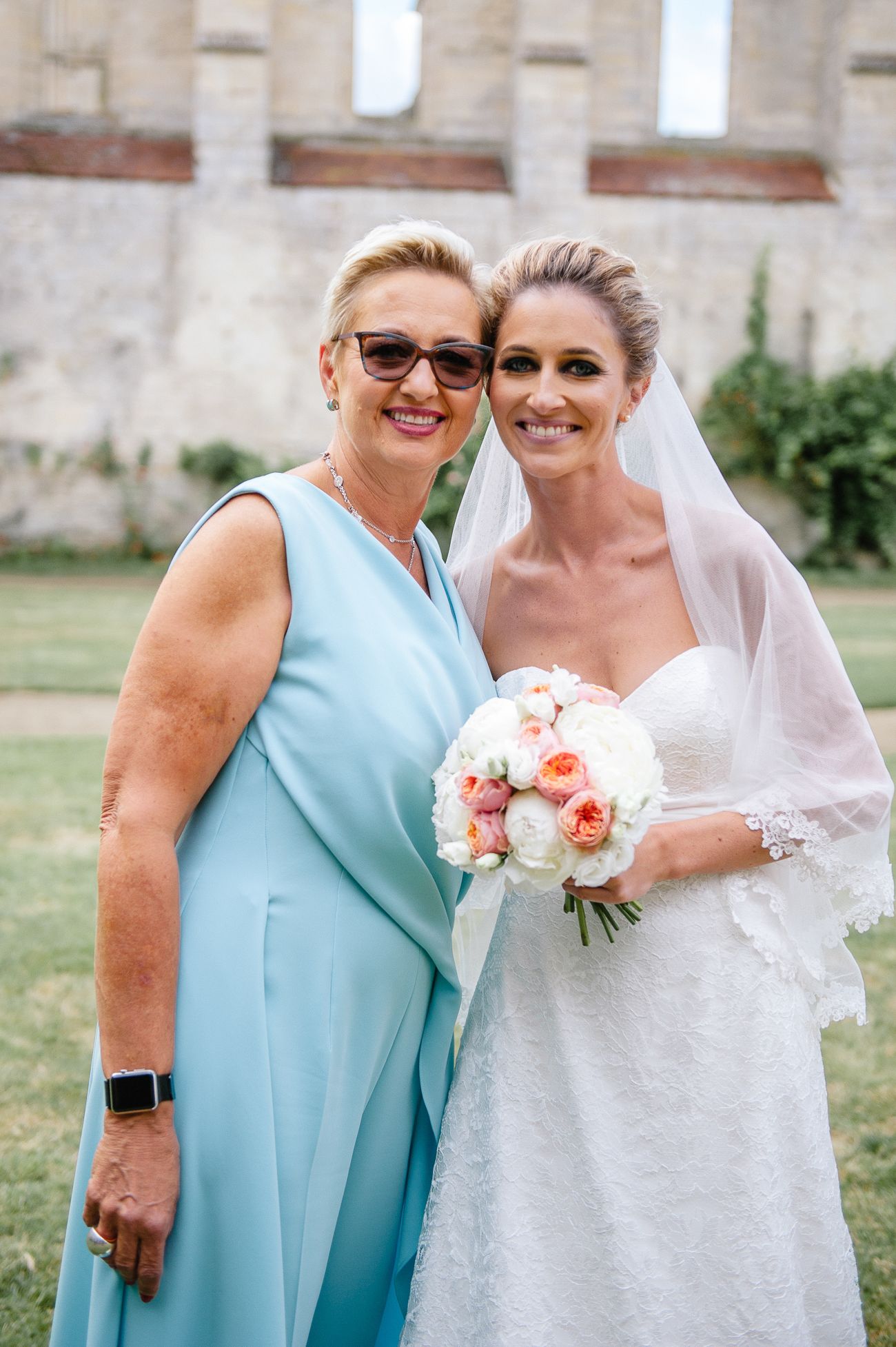 Smiling bride in lace gown holding peach bouquet poses with older woman in blue dress outdoors.