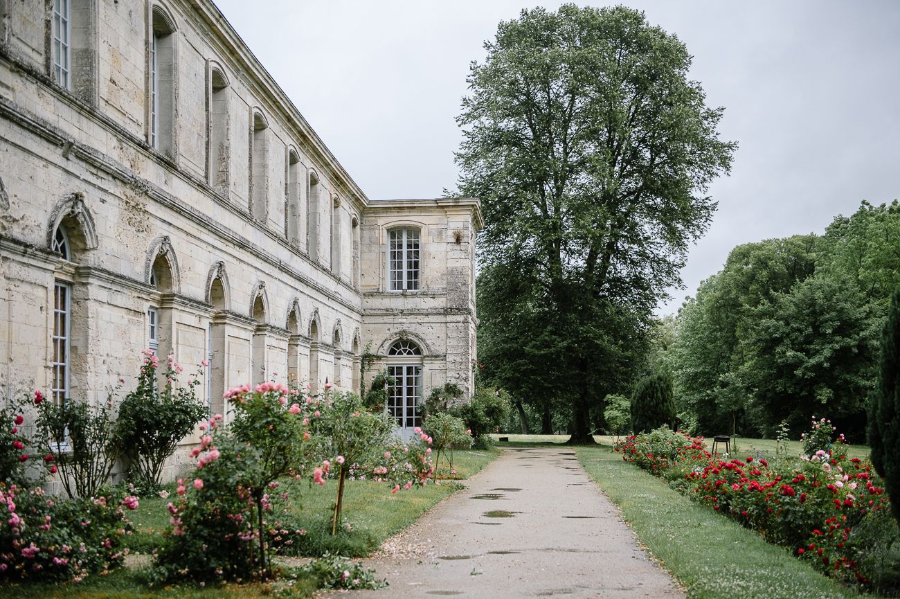 Historic French chateau with rose garden pathway, lush green trees, and elegant stone architecture on a cloudy day.