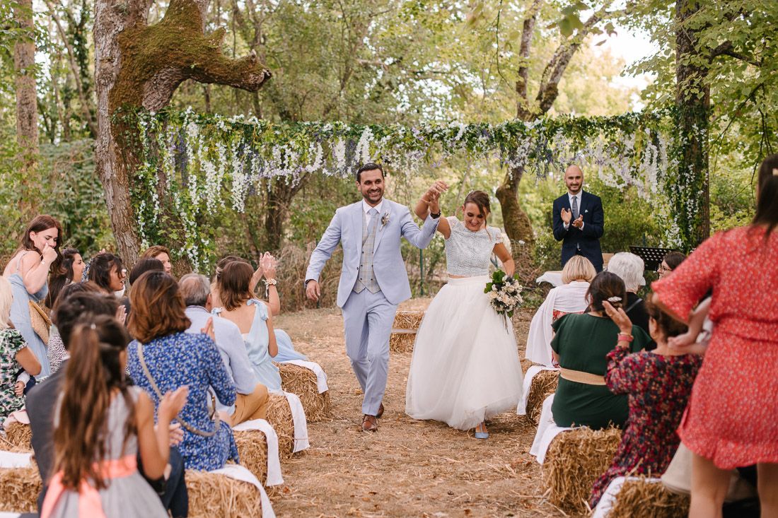 Newlyweds walk joyfully down aisle as guests applaud at rustic outdoor wedding with hay bale seating.
