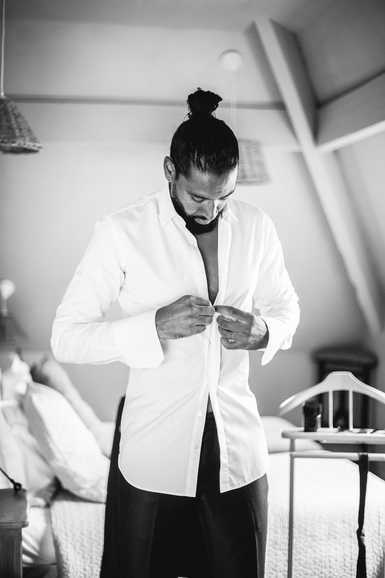 Man with bun hairstyle buttoning white dress shirt in bedroom, black and white portrait photo.