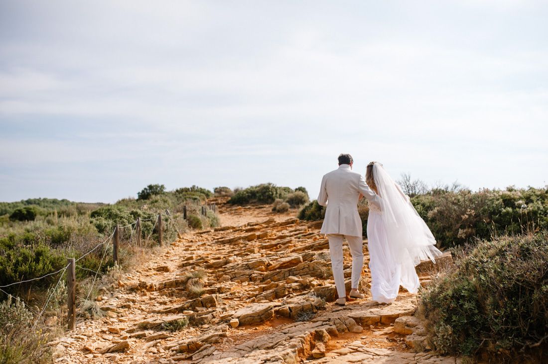 Bride and groom walking hand in hand along a rustic coastal path surrounded by green shrubs on their wedding day.