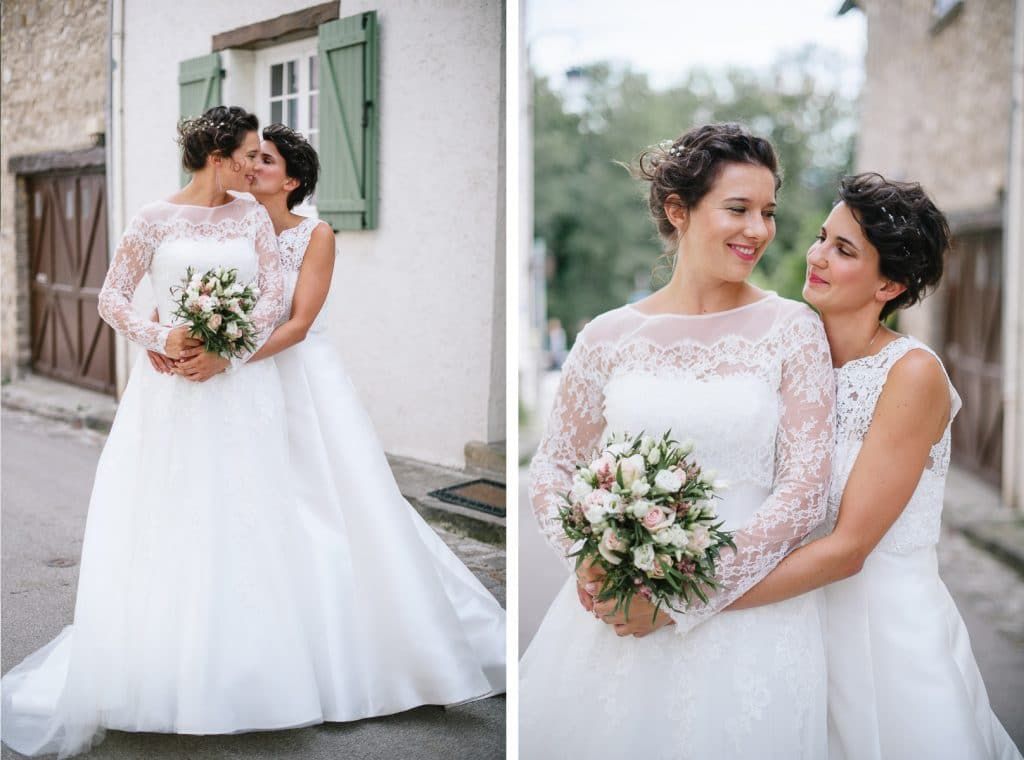 Two brides in white lace wedding gowns sharing a tender moment holding a floral bouquet outdoors.