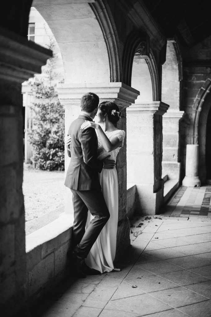 Black and white photo of newlyweds sharing an intimate moment under stone arches.