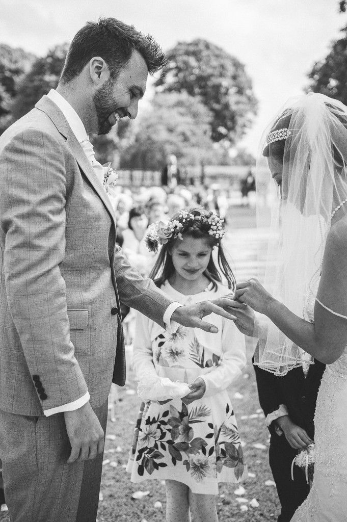 Smiling groom receives wedding ring from bride as adorable flower girl in floral dress watches during outdoor ceremony.