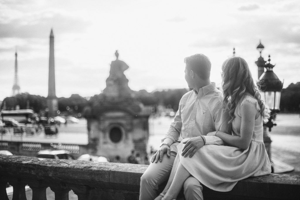 Black and white photo of a couple sitting together on a bridge ledge overlooking Paris landmarks.