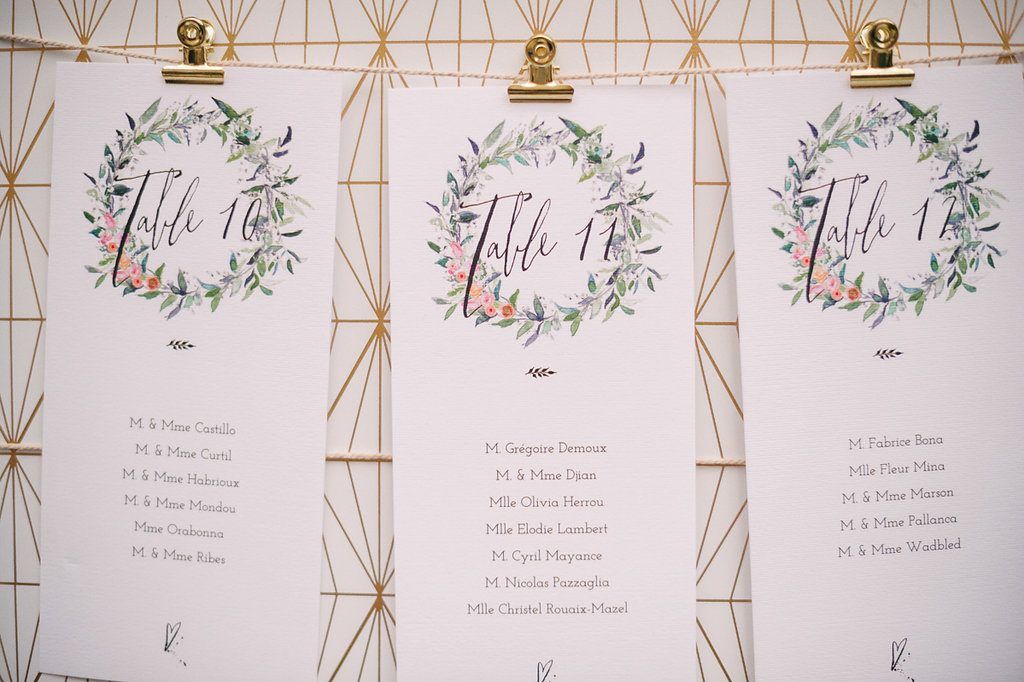 Elegant wedding seating chart cards with watercolor floral wreaths displayed on geometric gold frame backdrop.