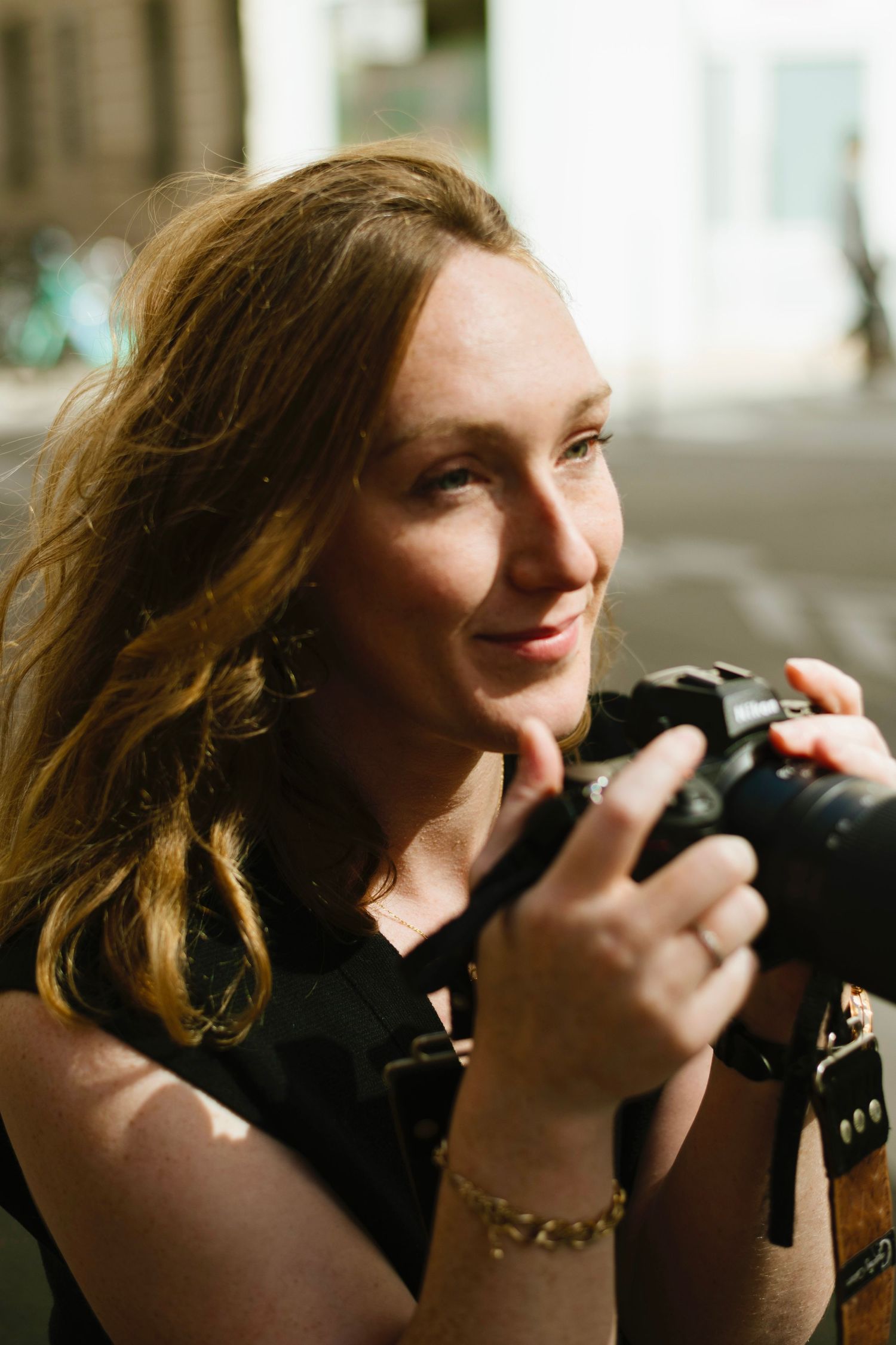 Young woman with wavy hair smiling while holding a DSLR camera outdoors in natural sunlight.