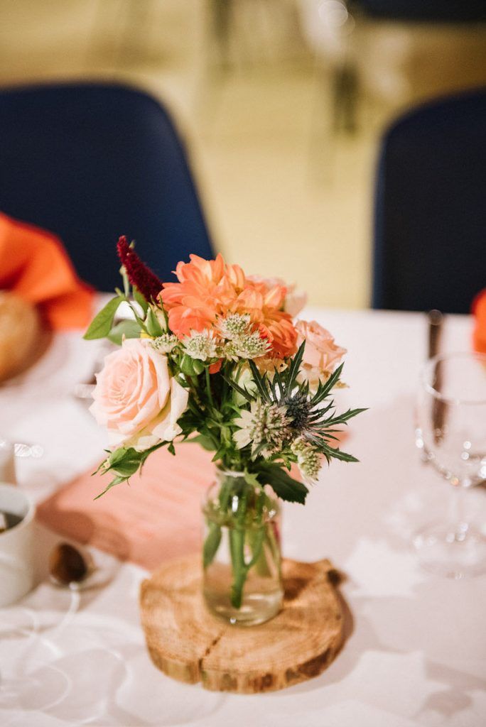 Wedding table centerpiece featuring peach dahlias, roses, and thistles in a glass jar on a wood slice.