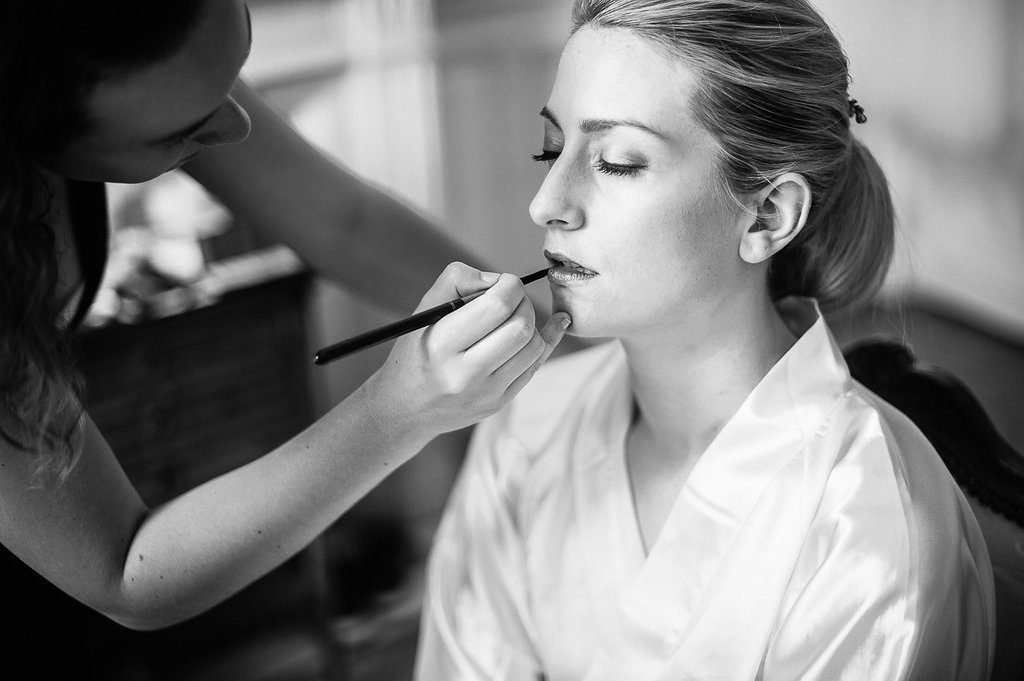 Black and white photo of makeup artist applying lipstick to a bride in a satin robe.
