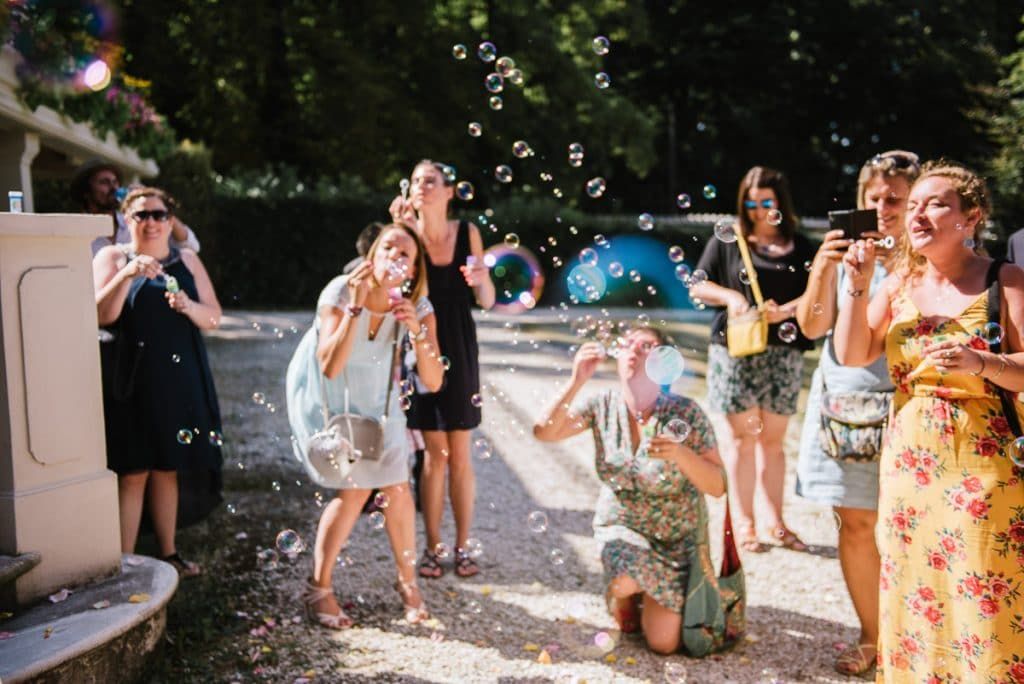 Wedding guests laughing and playing with soap bubbles outdoors in sunny garden during summer celebration.