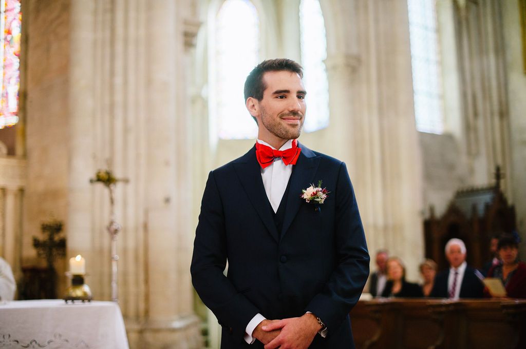 Groom in navy suit with red bow tie and boutonniere standing at church altar.