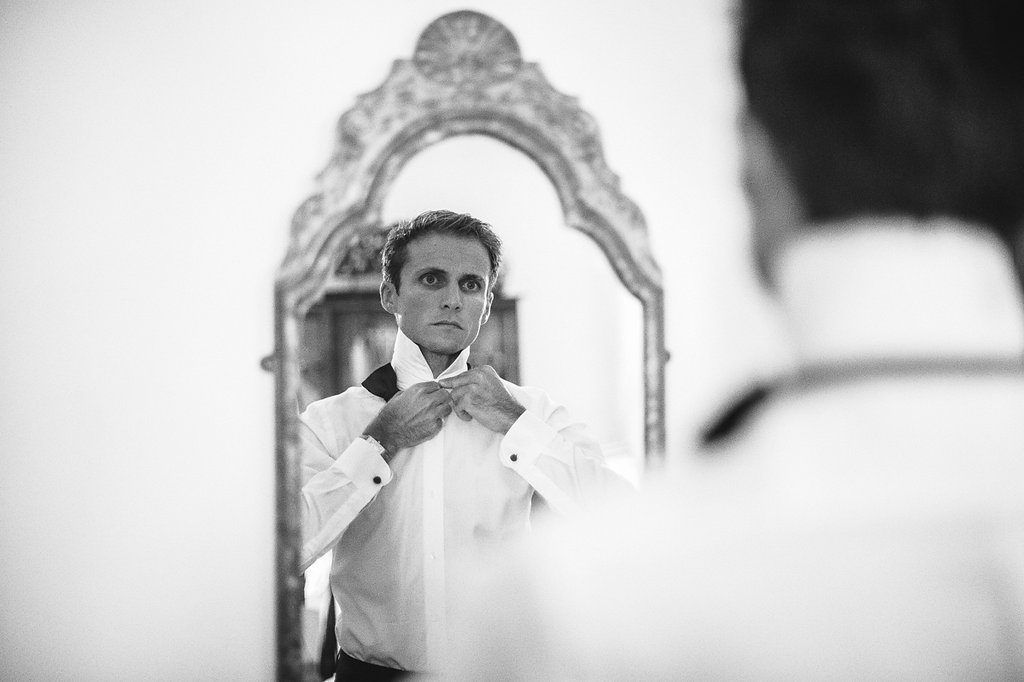 Groom adjusting bow tie while looking in ornate vintage mirror before wedding.