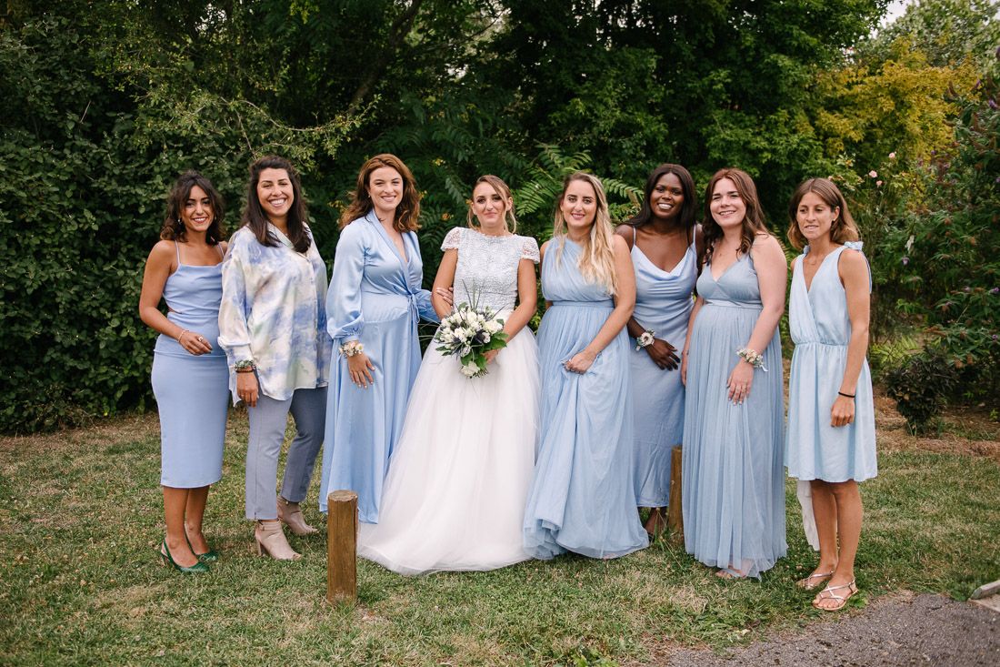 Bride in white gown poses with six bridesmaids in light blue dresses outdoors on a sunny day.