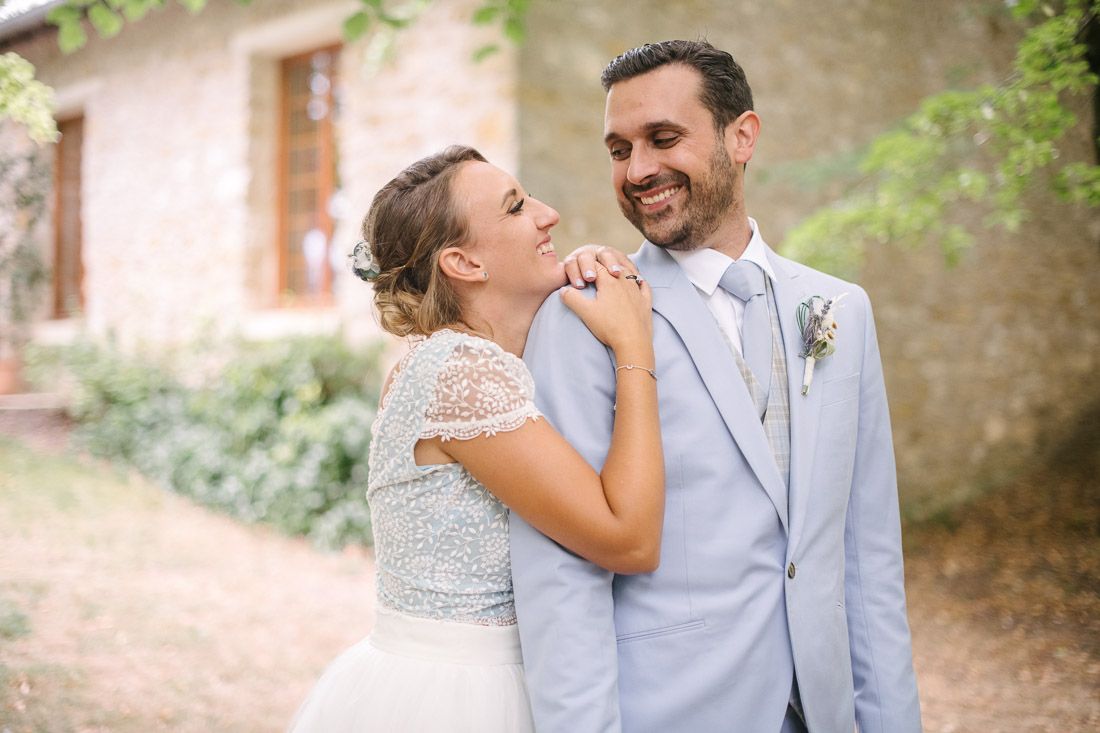 Bride in lace crop top wedding dress embraces smiling groom in light blue suit outdoors at rustic venue.