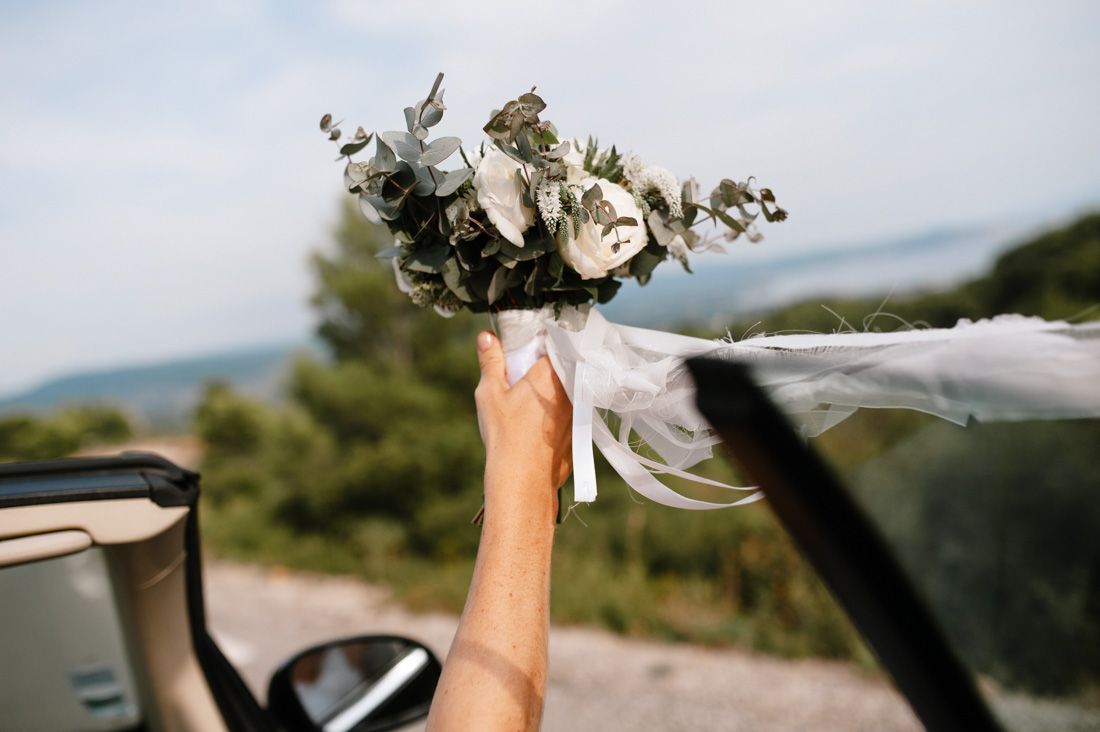 A hand holds a white wedding bouquet with eucalyptus out of a convertible car window against a scenic backdrop.