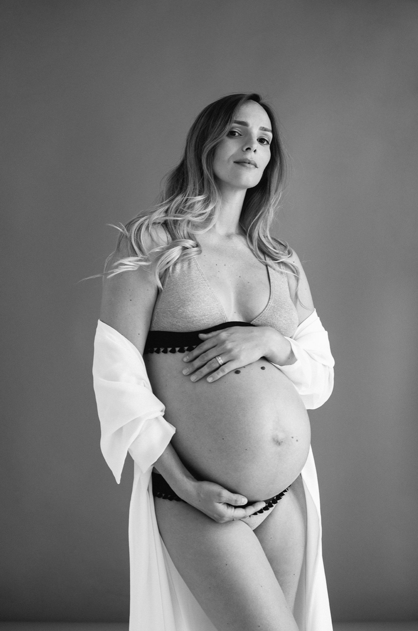 Black and white maternity photo of pregnant woman in lingerie with white robe draping her shoulders.
