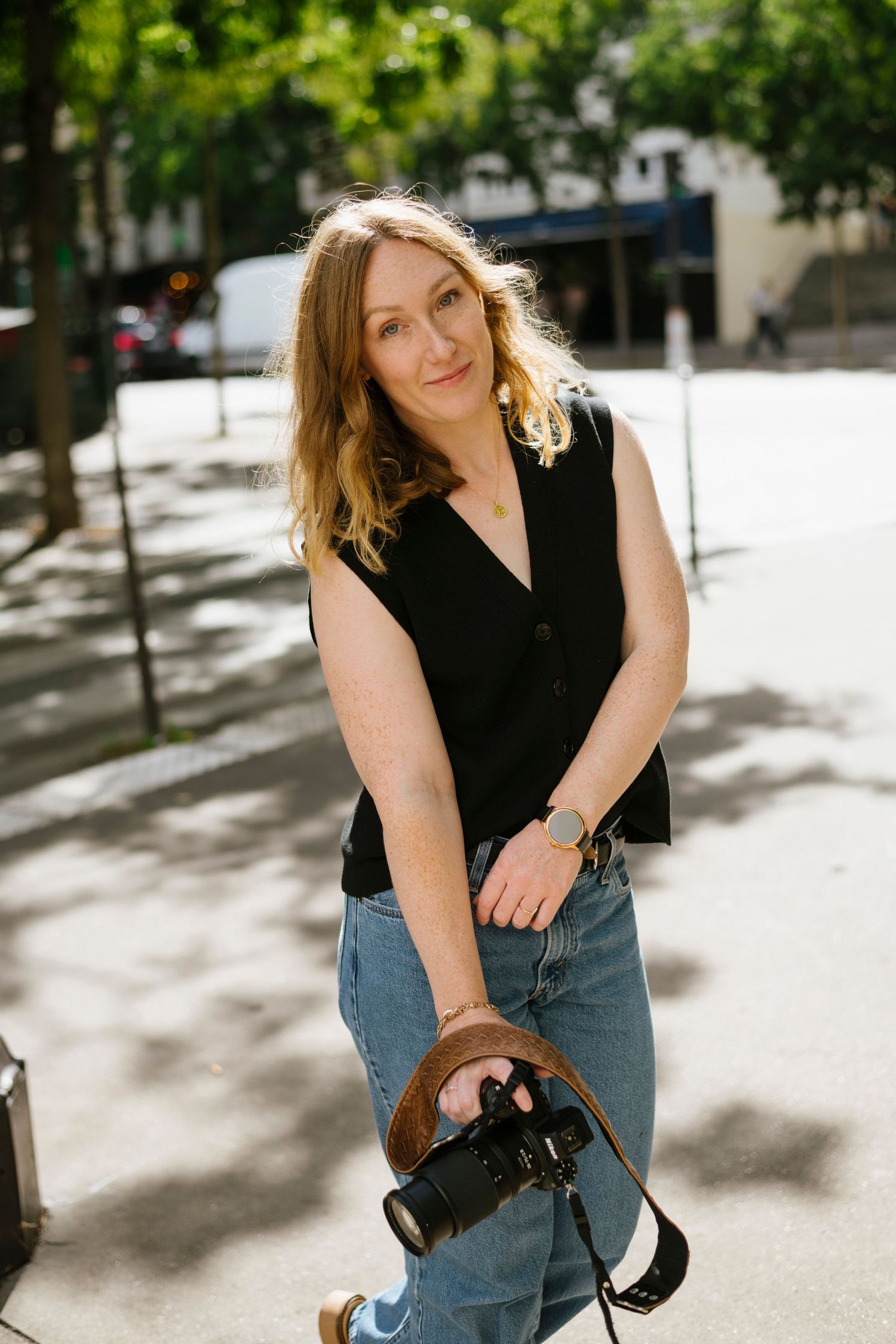 Woman with blonde wavy hair holding a camera while posing outdoors on a sunny tree-lined street.