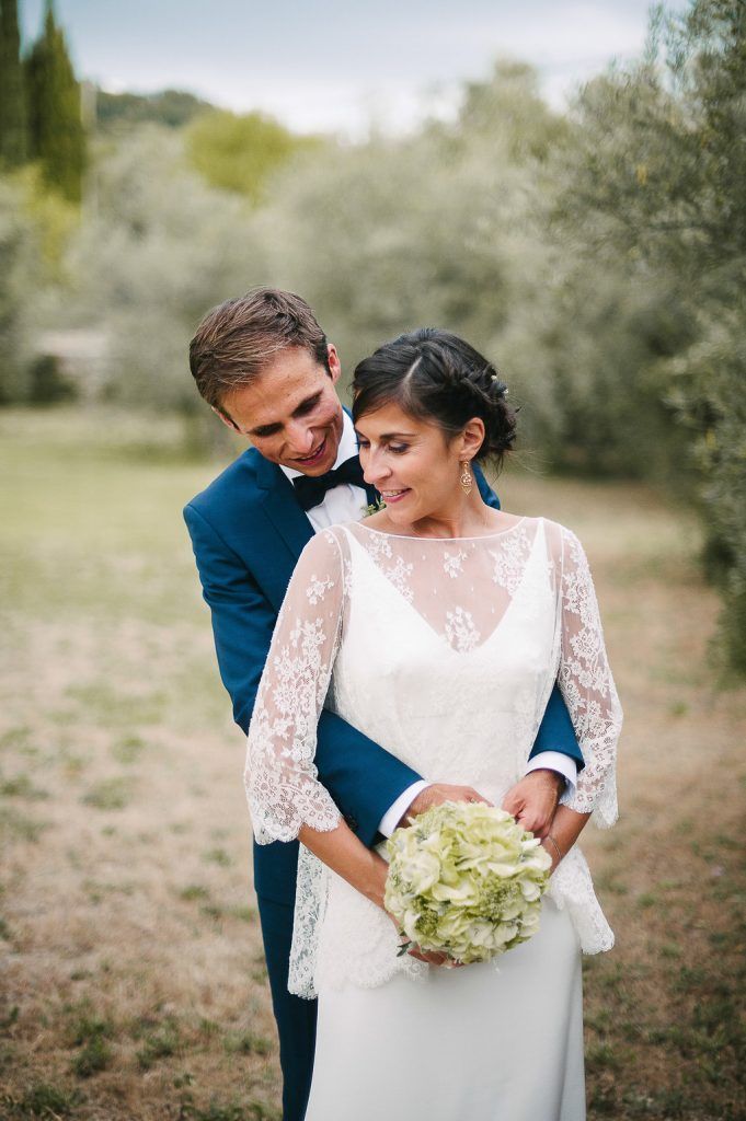 Groom in navy suit embraces smiling bride in lace wedding dress holding green hydrangea bouquet outdoors.
