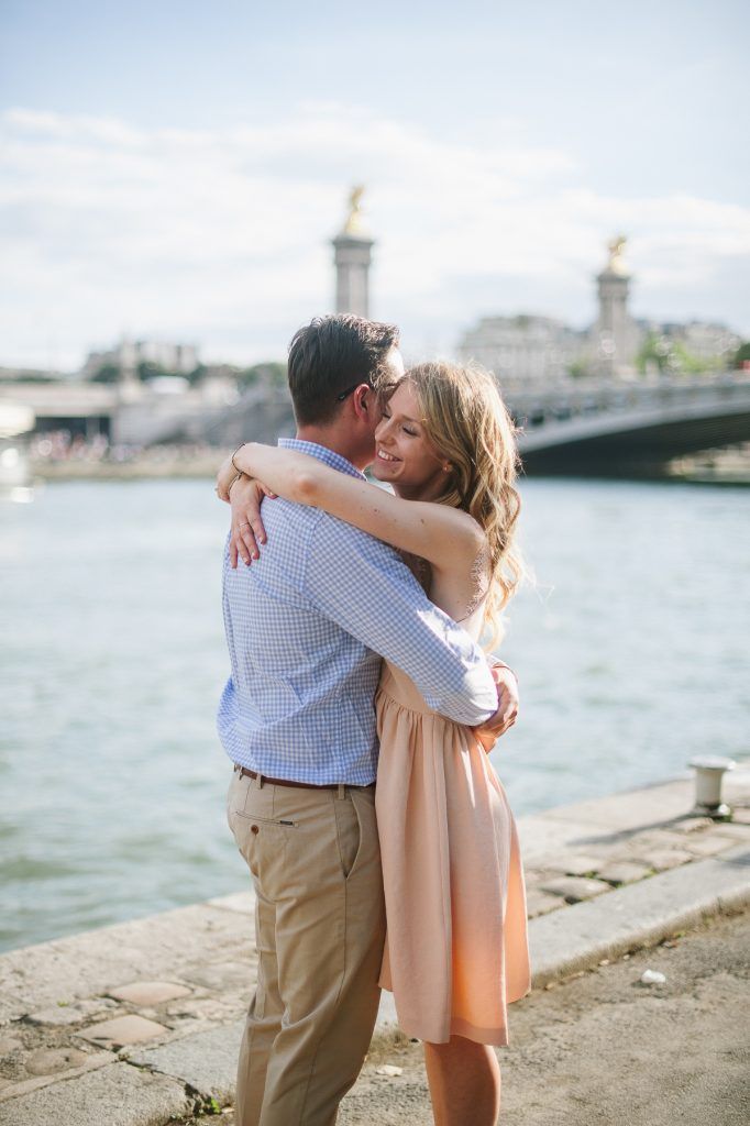 Couple hugging lovingly by the Seine River with Pont Alexandre III bridge in background.