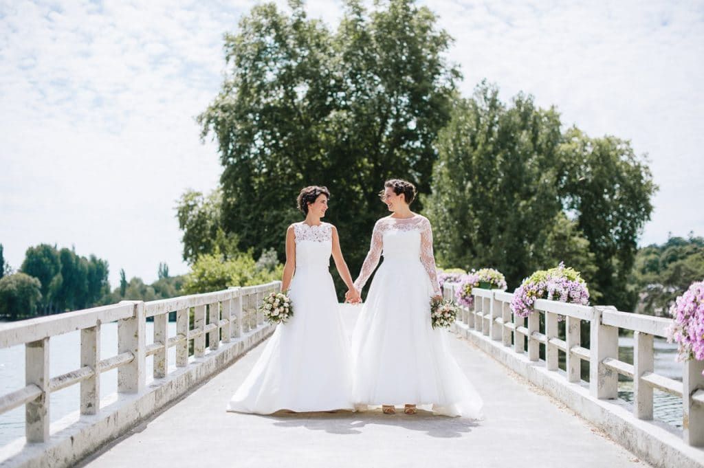 Two brides in white wedding gowns holding hands walking on a bridge.