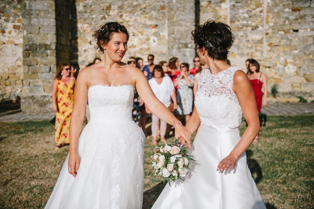 Two brides in white gowns hold hands and smile joyfully during their outdoor wedding ceremony.