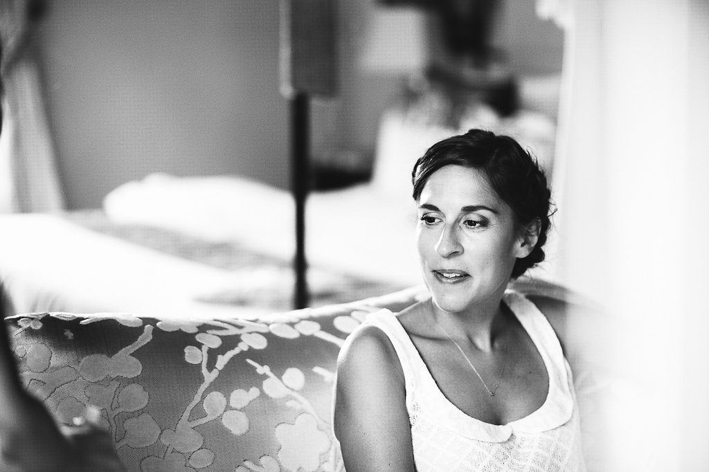 Black and white portrait of brunette bride in white dress sitting near bright window.