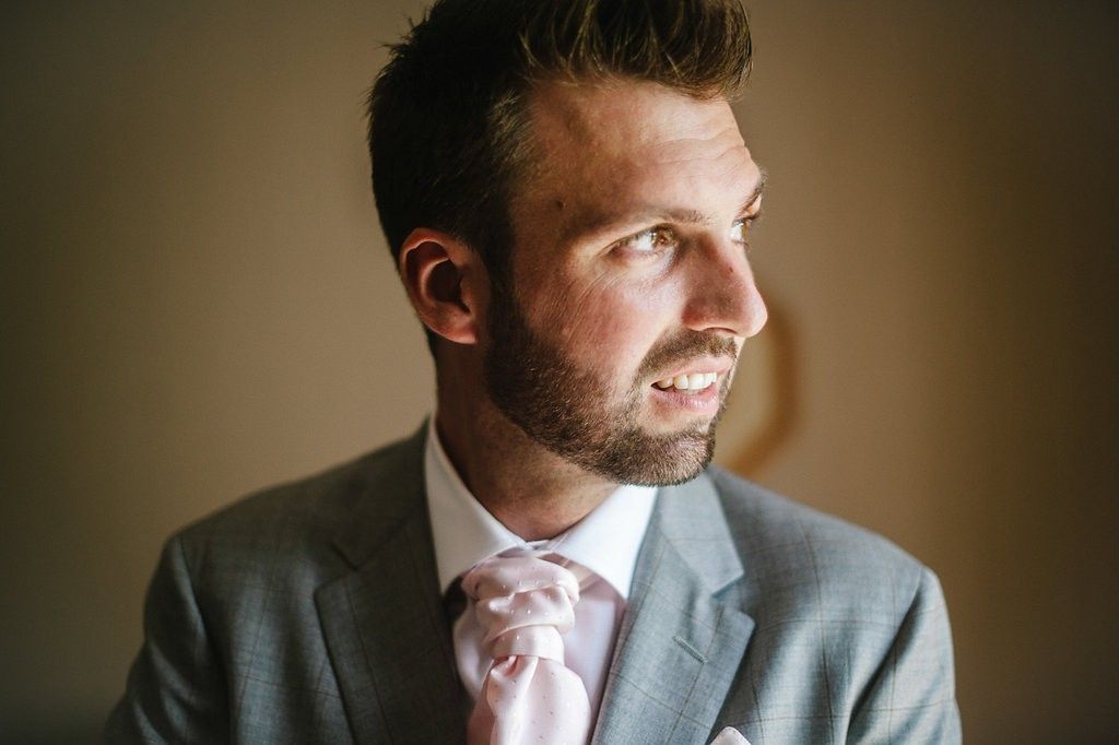 Handsome bearded groom in gray suit and pink tie smiling on wedding day.