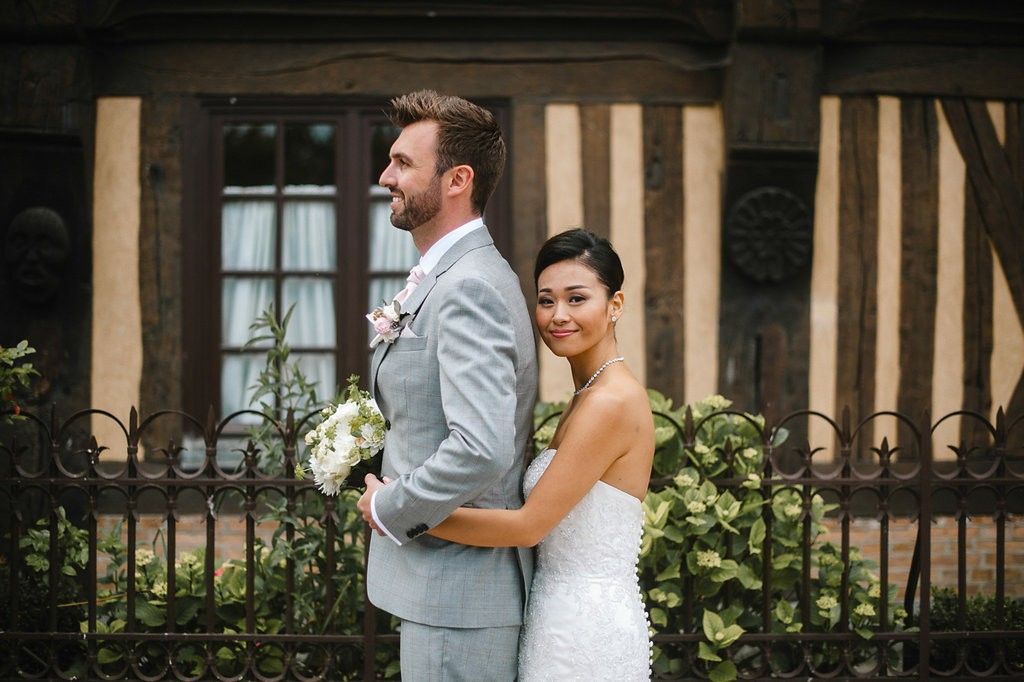 Bride and groom pose back-to-back in front of a Tudor-style building, bride in white gown holding bouquet.