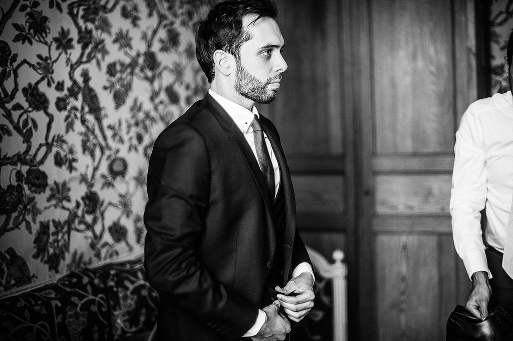Groom in black suit standing in ornate wallpapered room during wedding day preparations.