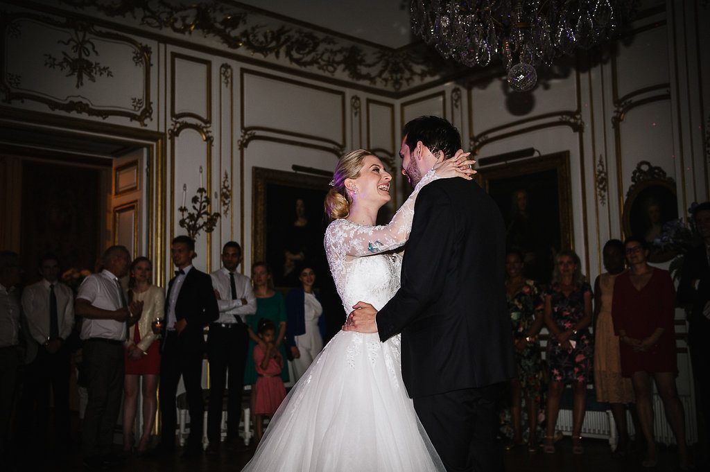 Newlyweds share their first dance in ornate ballroom, bride wearing lace gown at French chateau wedding.