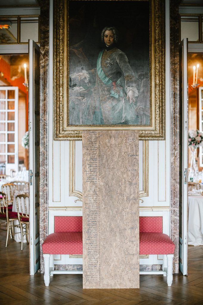 Elegant wedding seating chart displayed on marble stand beneath ornate portrait in grand French chateau ballroom.