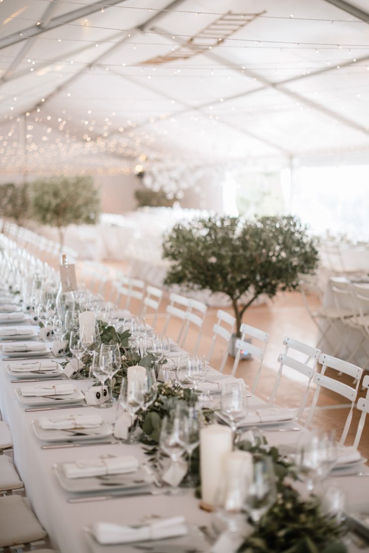 Elegant wedding reception tent with long tables, white chairs, greenery runners, candles, and fairy lights overhead.