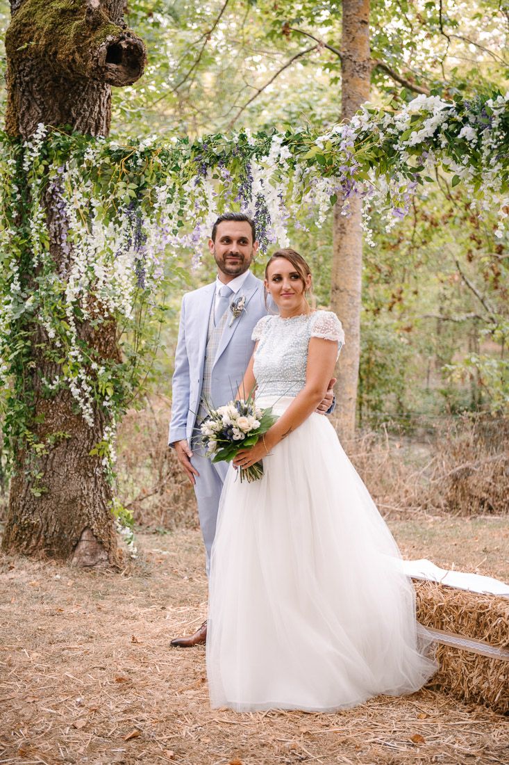 Bride and groom pose under floral arch with wisteria and greenery in woodland wedding setting.