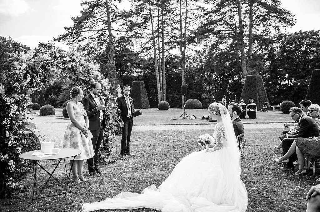 Black and white photo of outdoor garden wedding ceremony with bride, officiant, and seated guests among sculpted hedges.