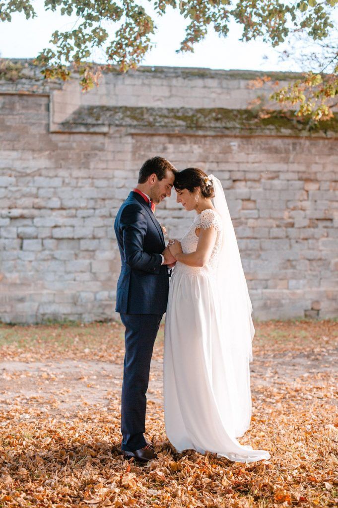 Bride and groom hold hands tenderly in autumn leaves before rustic stone garden wall.